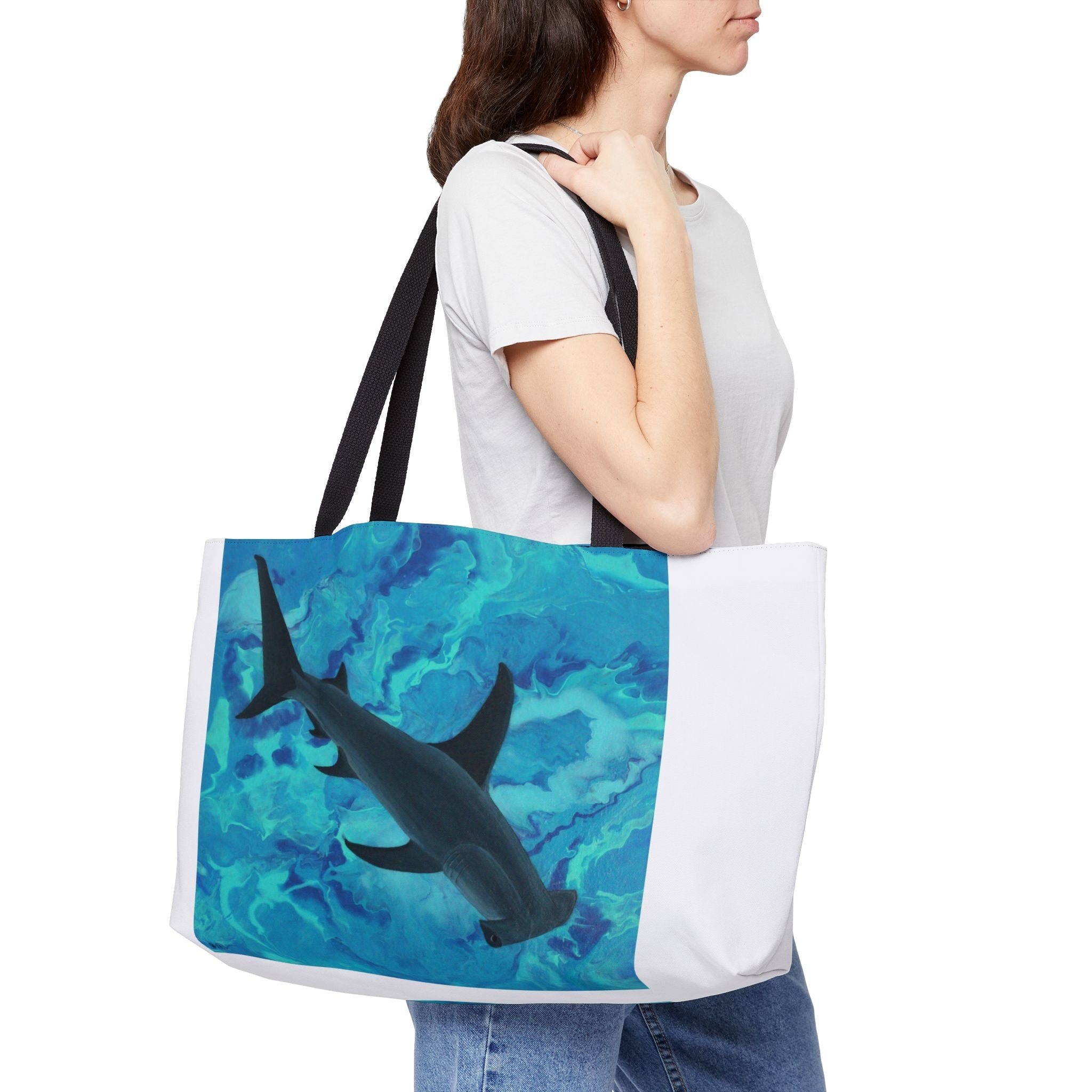 women in white shirt and jeans holding a weekender bag with black handles white bag with image of hammerhead shark swimming in turquoise sea
