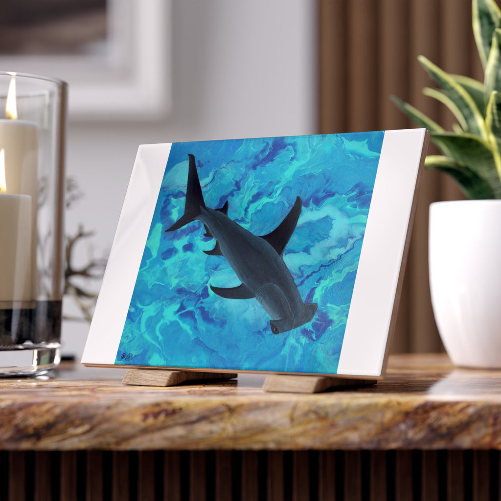 Tile with a hammerhead shark on a blue wave background, placed on a wooden surface with candles and a plant in the background.