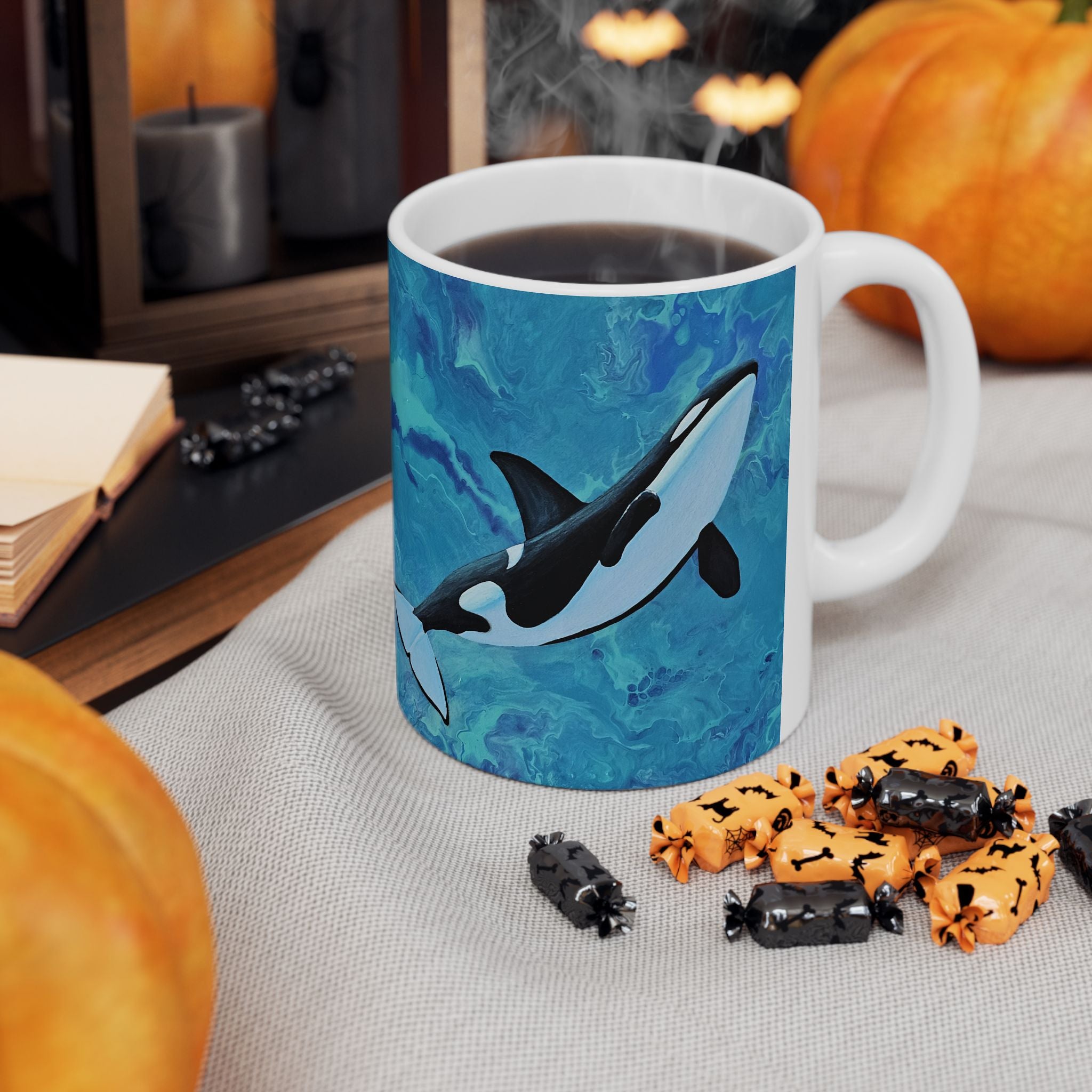 Mug with orca whale design on a table with Halloween-themed treats and pumpkins.