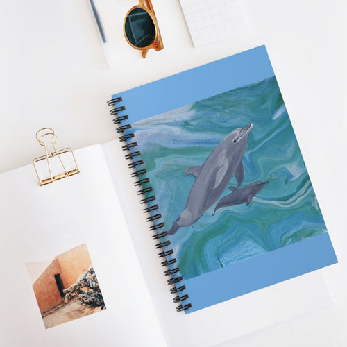 Notebook with dolphin design on cover, placed on a white surface with sunglasses and a photo clip.
