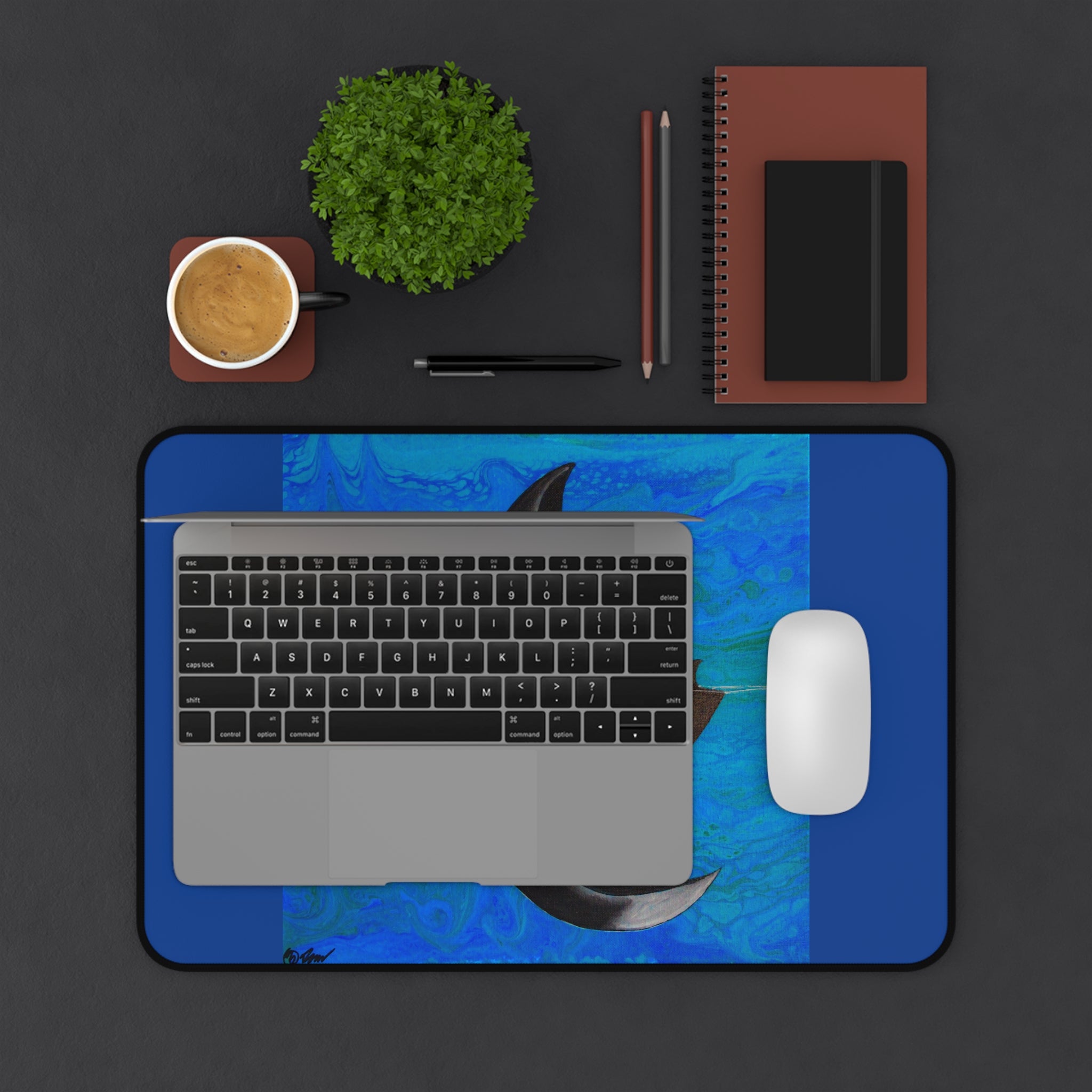 Laptop on a blue desk pad with a cup, plant, and stationery items on a dark surface