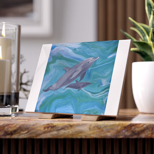 Tile with small painting of two dolphins on a stand with candles and a plant in the background