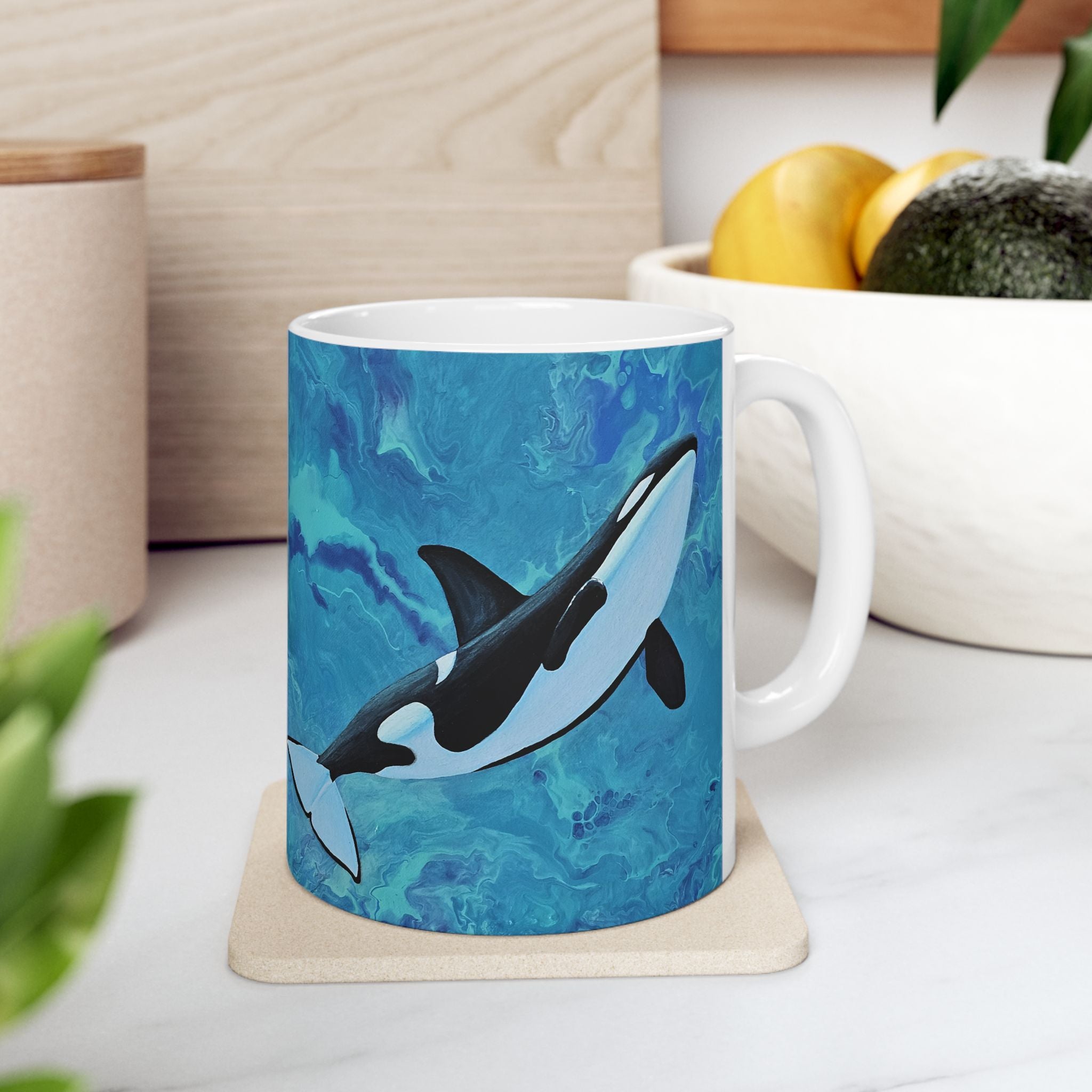 Mug with an orca whale design on a coaster, placed on a surface with a plant and container in the background.