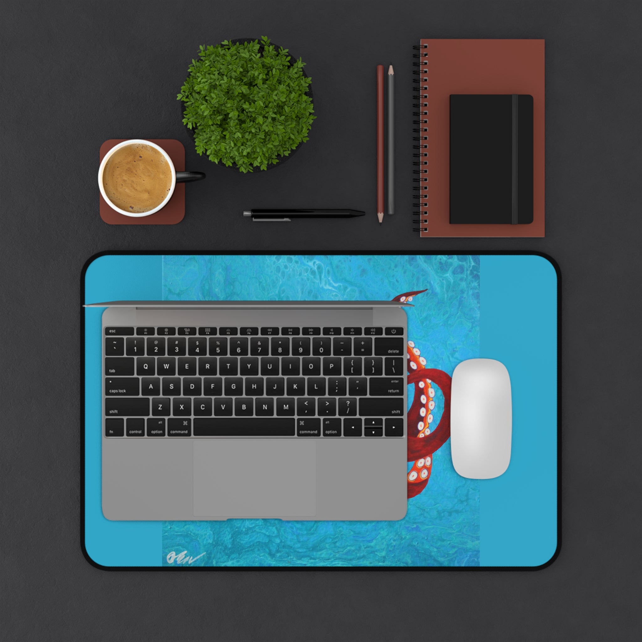 Laptop on a blue desk mat with a coffee cup, pen, and notebook on a dark surface