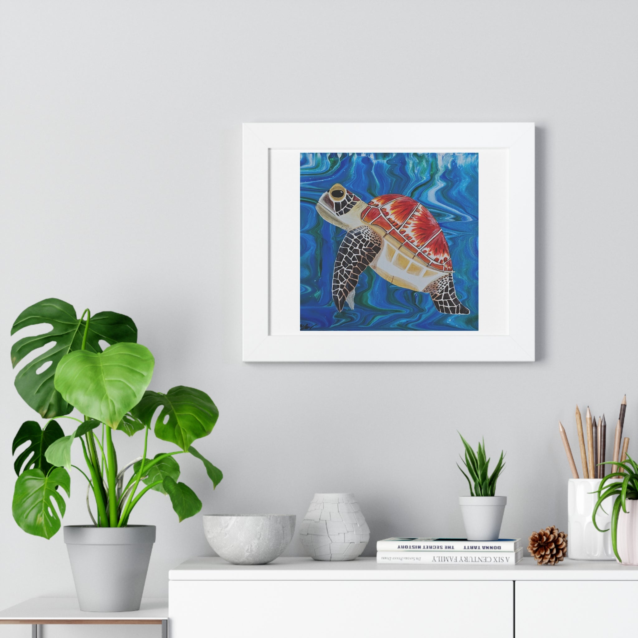 Framed artwork of a turtle in water on a wall above a white cabinet with plants.