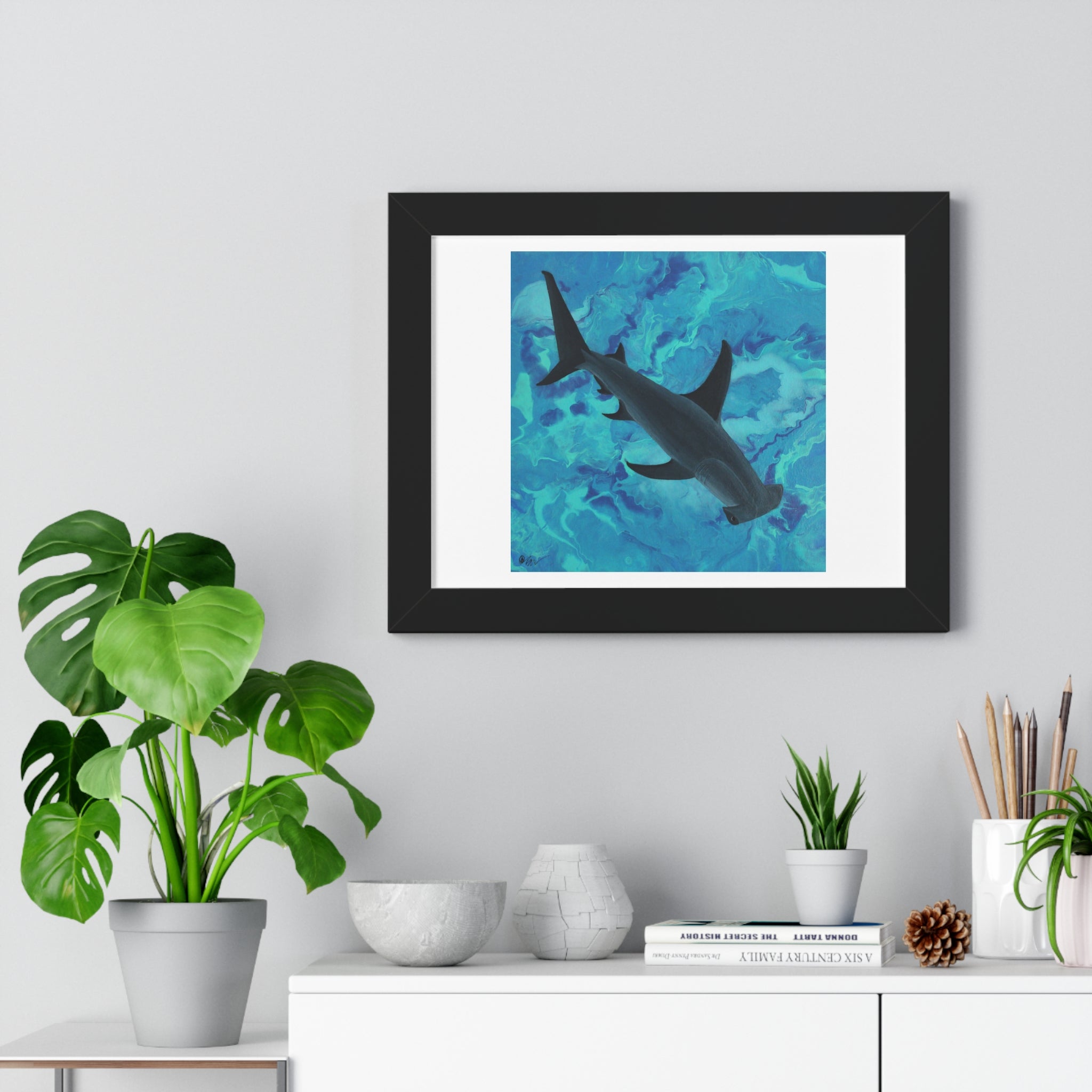 Framed artwork of a shark on a blue background in a home setting with plants and decor.