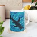 Mug with a shark design on a coaster, placed on a surface with a plant and container in the background.