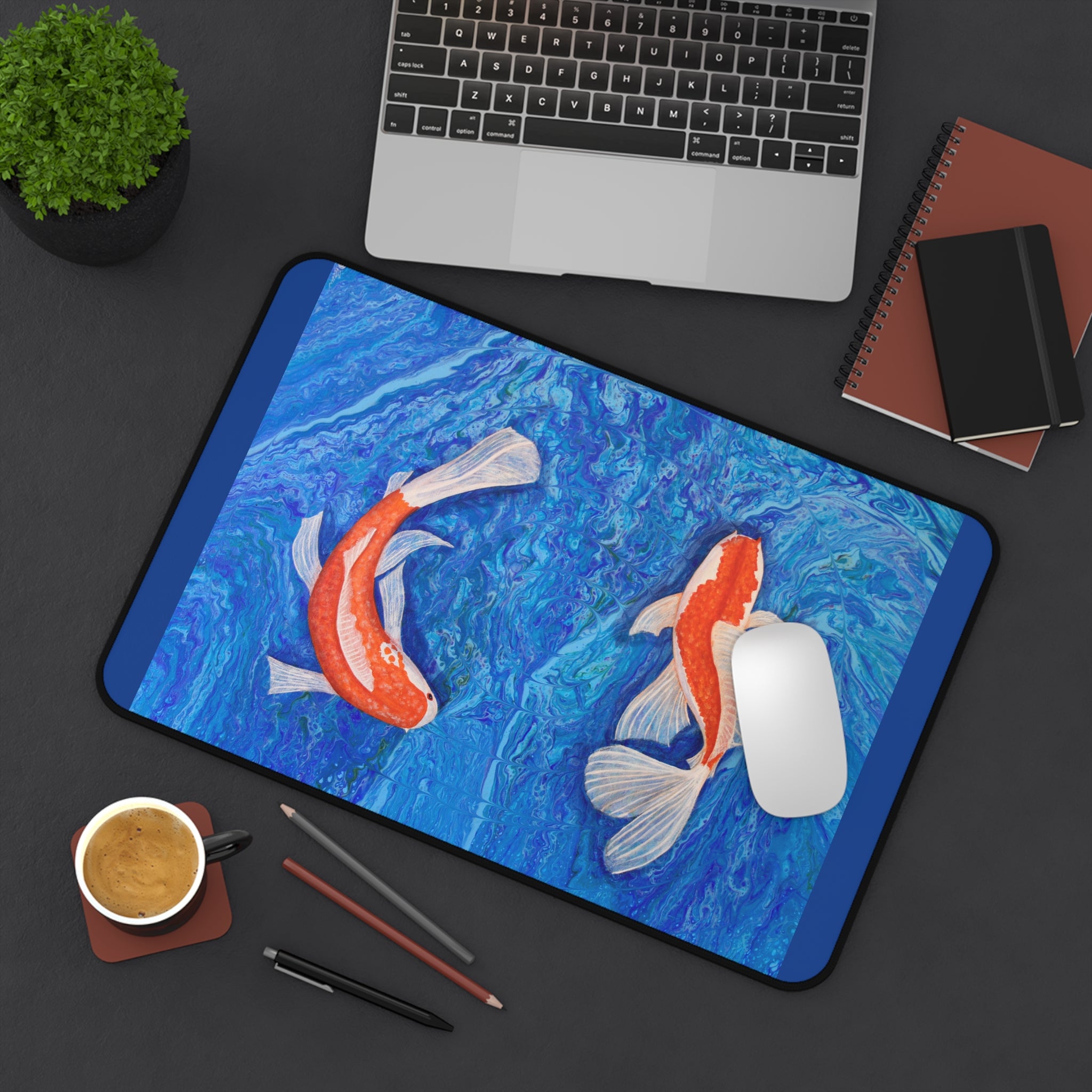 Desk setup with a desk pad featuring koi fish design, laptop, notebook, and coffee cup.