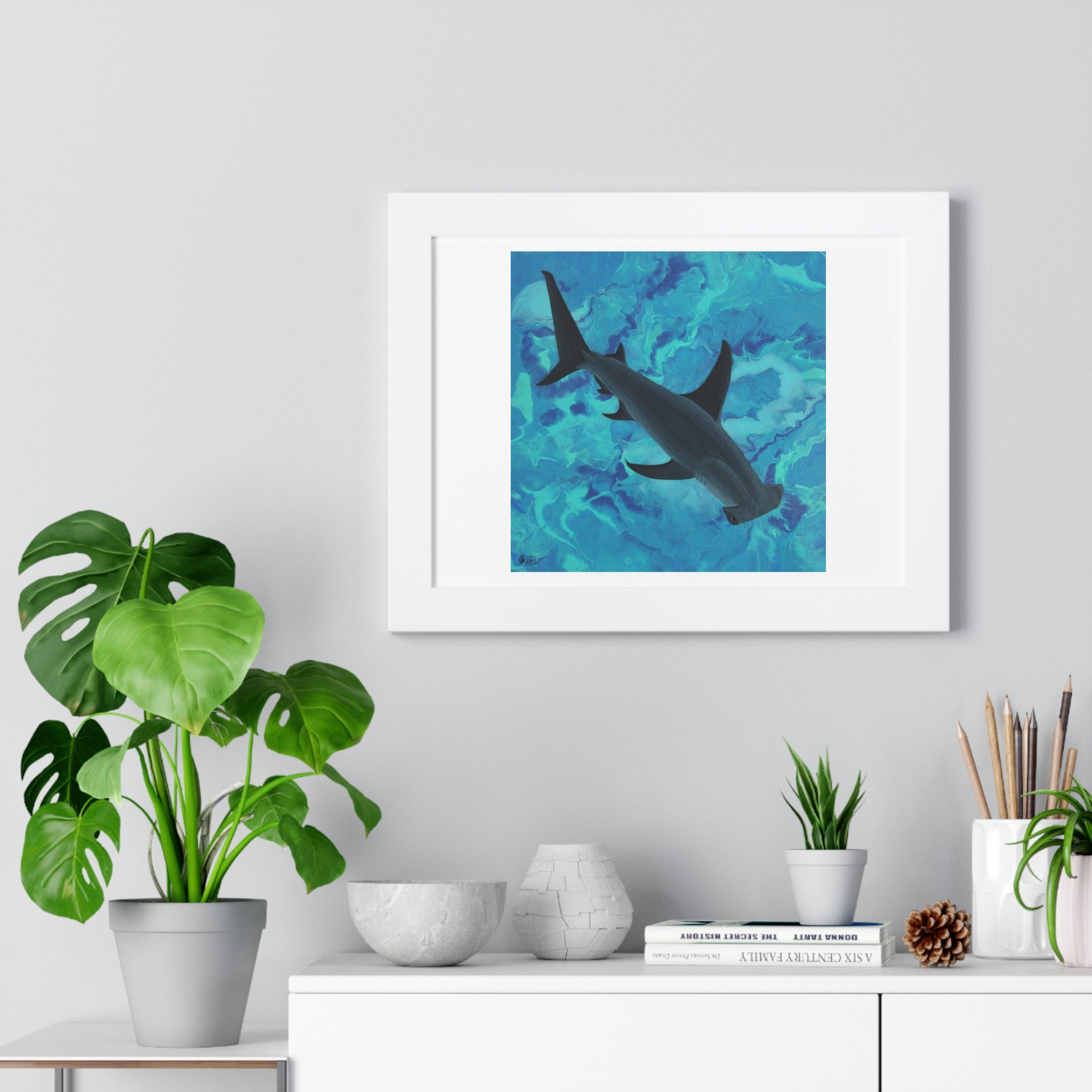 Framed artwork of a shark in water on a wall above a desk with plants and decor.