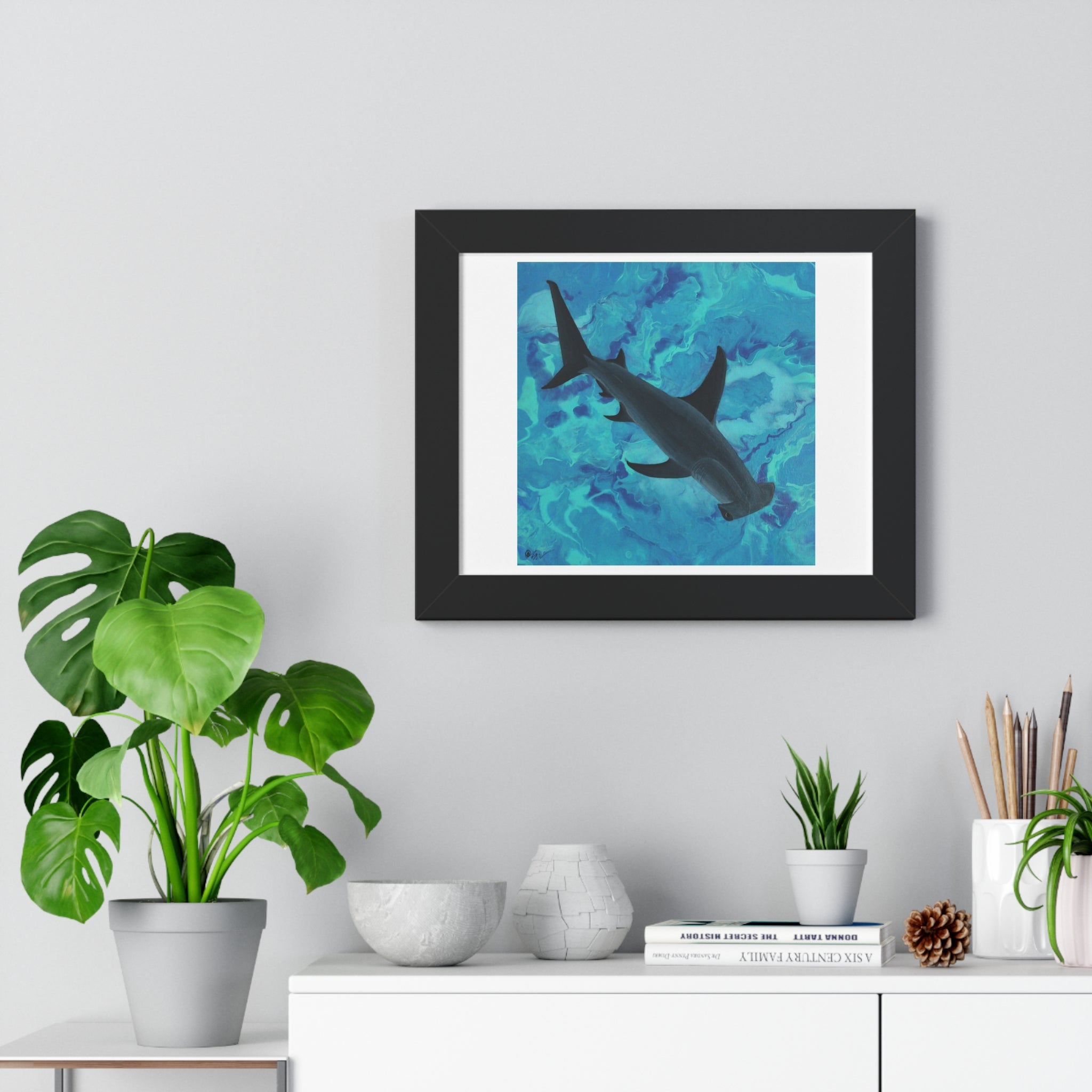 Framed artwork of a shark in water on a wall above a desk with plants and decor.