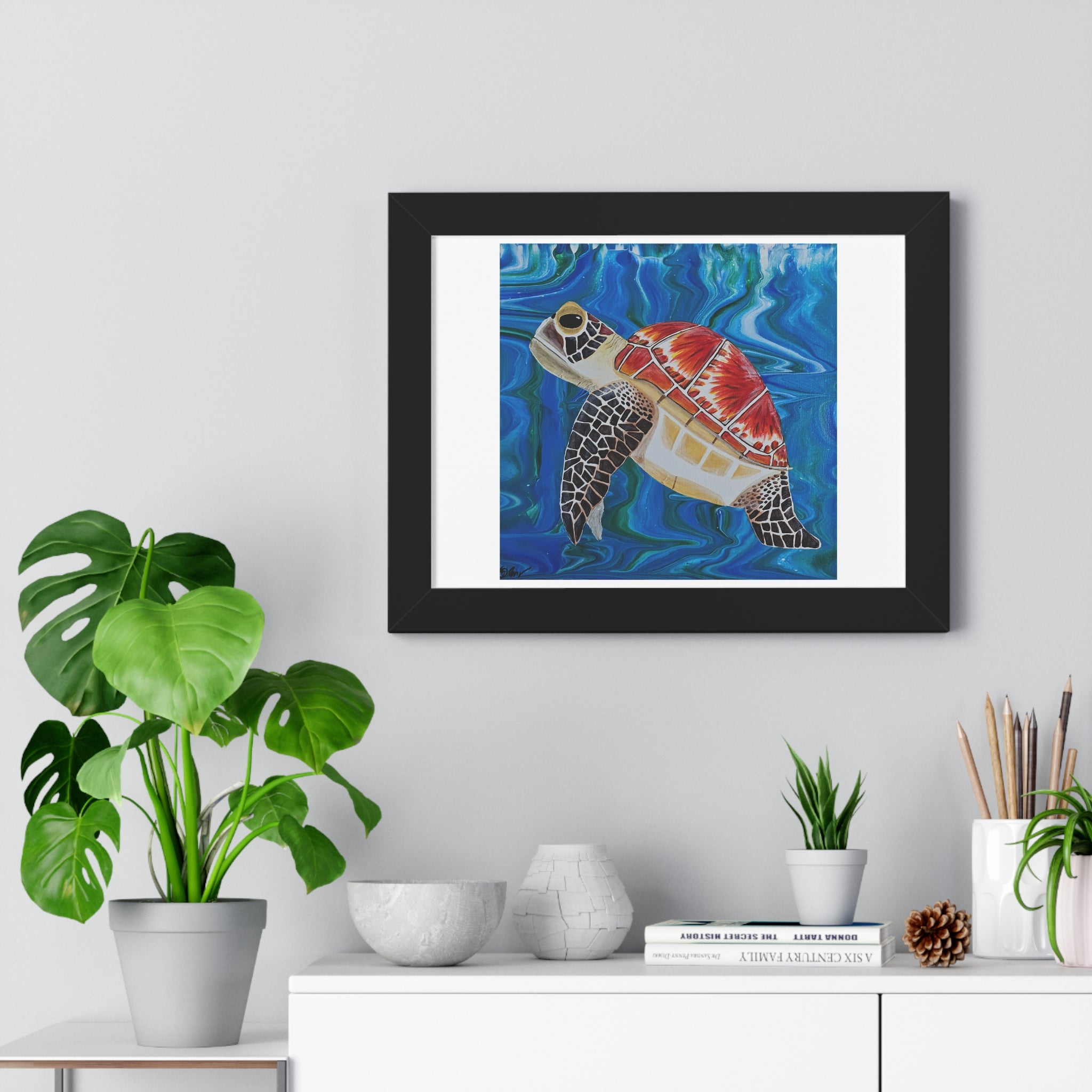Framed artwork of a turtle in an underwater scene on a wall above a white cabinet with plants.