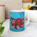 Mug with a red octopus design on a blue background, placed on a white surface with a plant and container in the background.