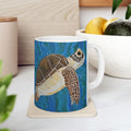 Mug with a turtle design on a coaster, placed on a surface with a plant and container in the background.
