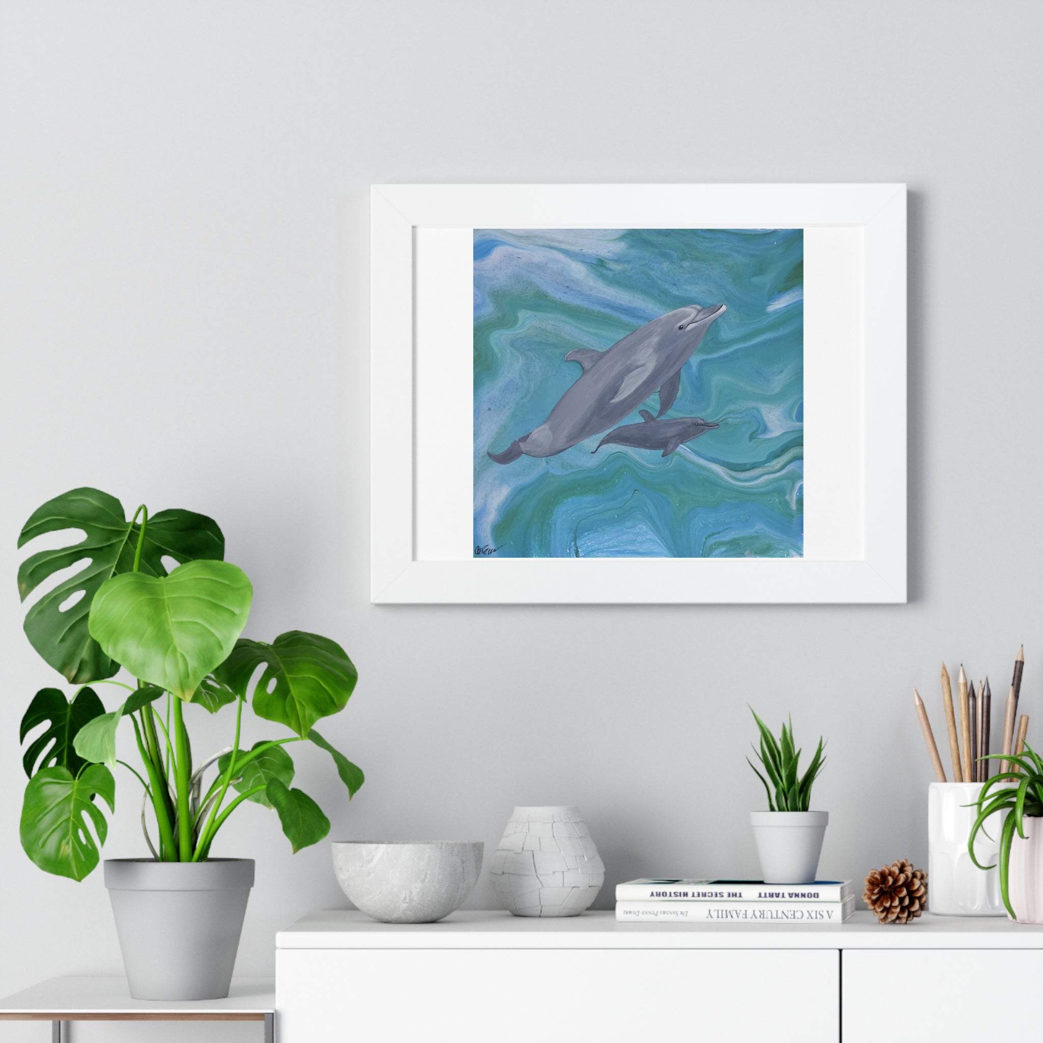 Framed artwork of dolphins in water on a wall above a desk with plants and decor.