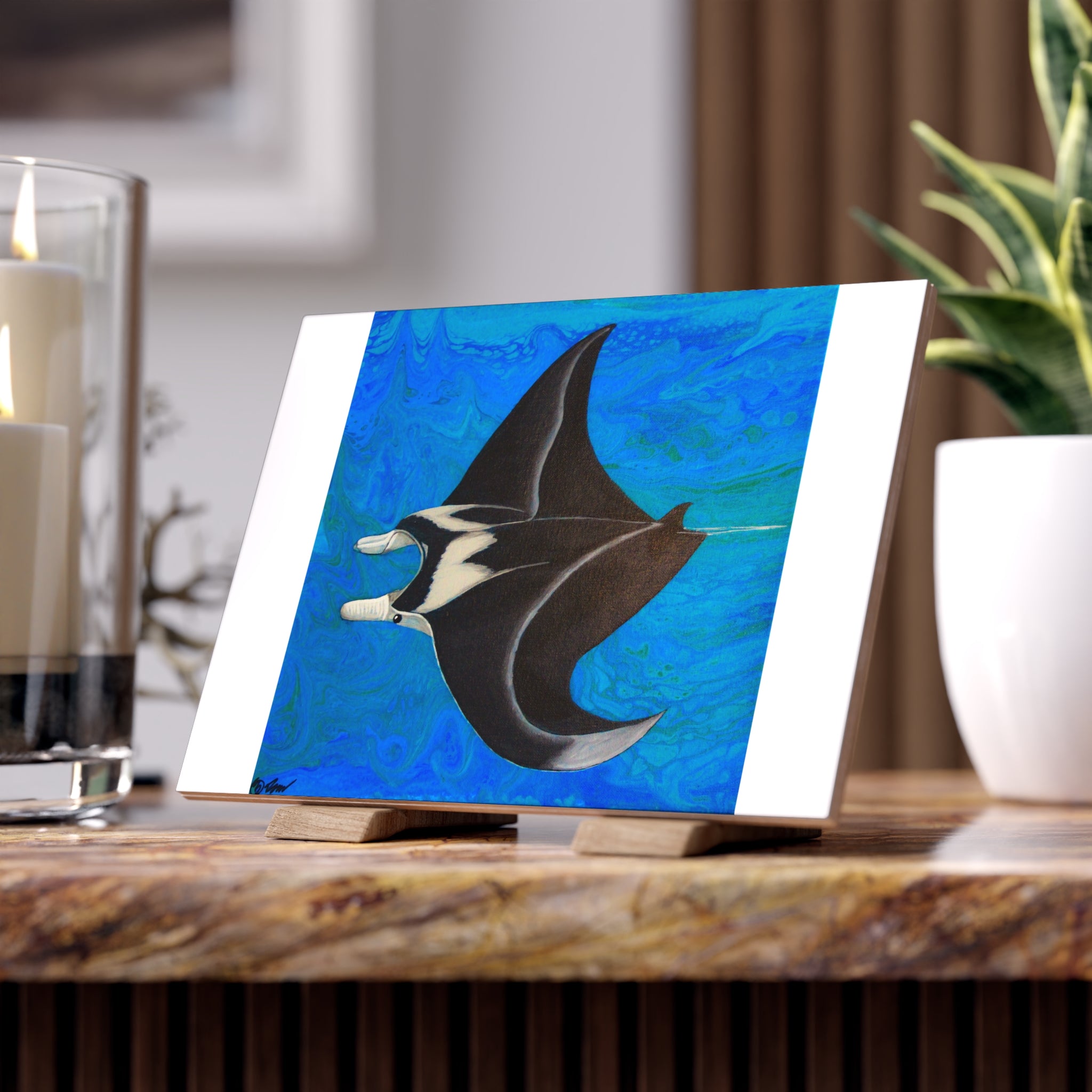 Artistic depiction of a manta ray on a blue background tile, placed on a wooden surface with candles and a plant in the background.