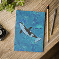Notebook with an orca whale design on a wooden surface with a plant and stationery items.
