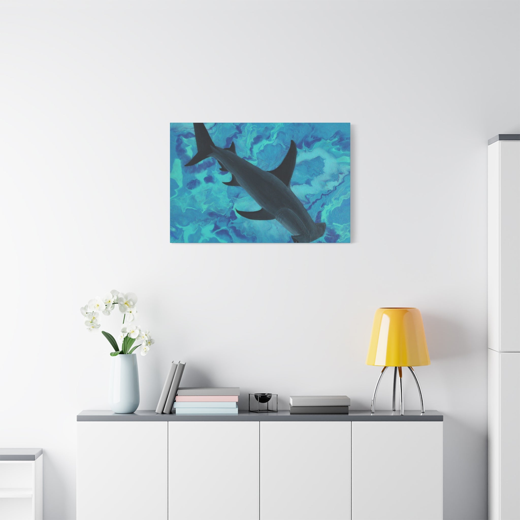 Shark painting on a wall above a white cabinet with a yellow lamp and white flowers.