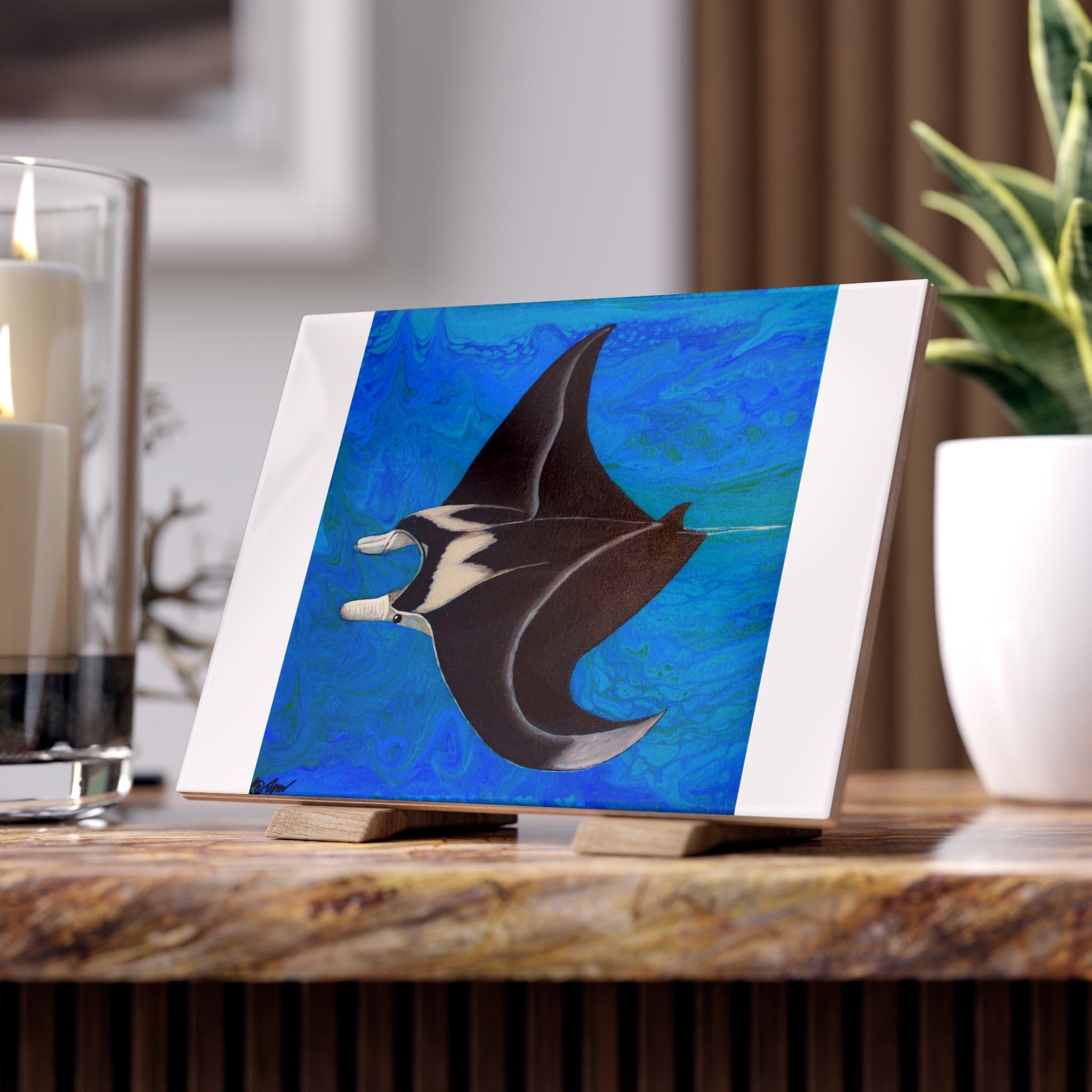 Artistic depiction of a manta ray on a blue background tile, placed on a wooden surface with candles and a plant in the background.