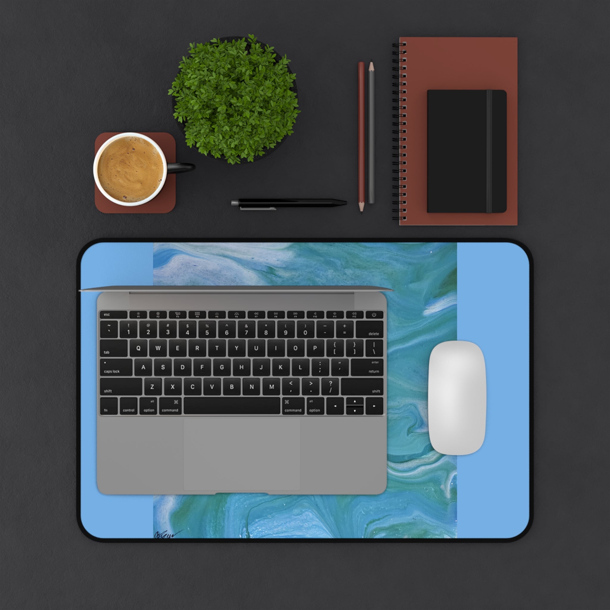 Laptop on a blue desk pad with a coffee cup, plant, and stationery items on a dark surface